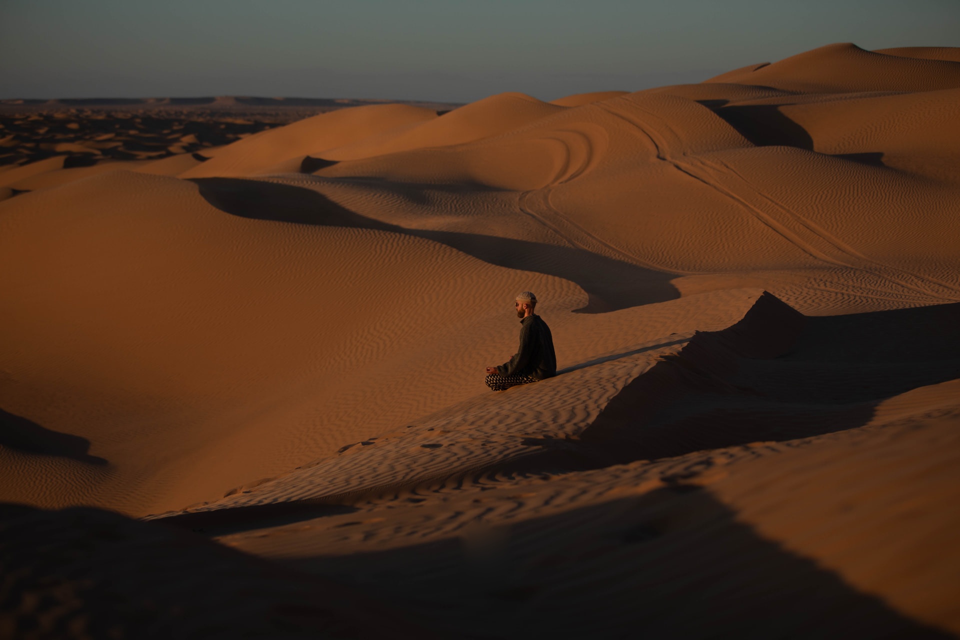 A meditating figure seated alone on a vast desert dune at sunset